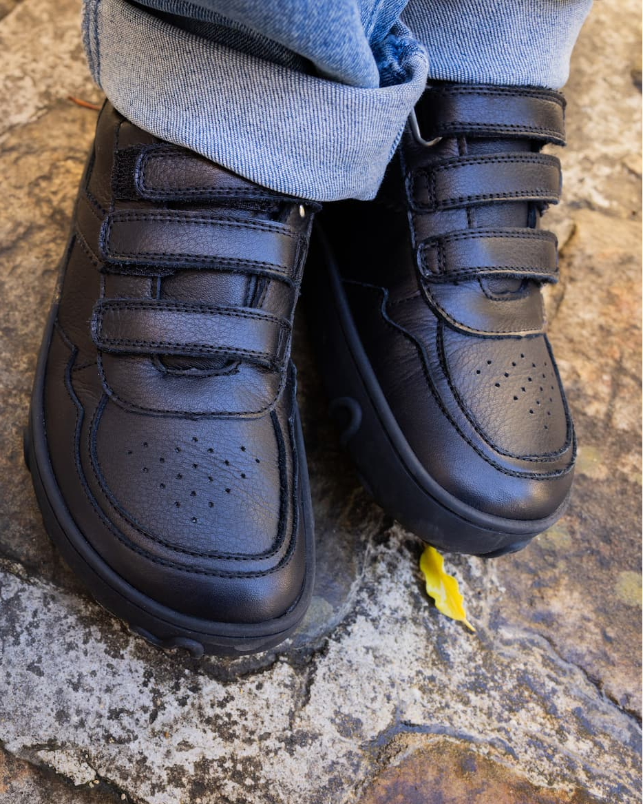 Black leather toddler barefoot sneakers with three hook-and-loop straps, shown on a child's feet with cuffed blue jeans standing on a stone surface.