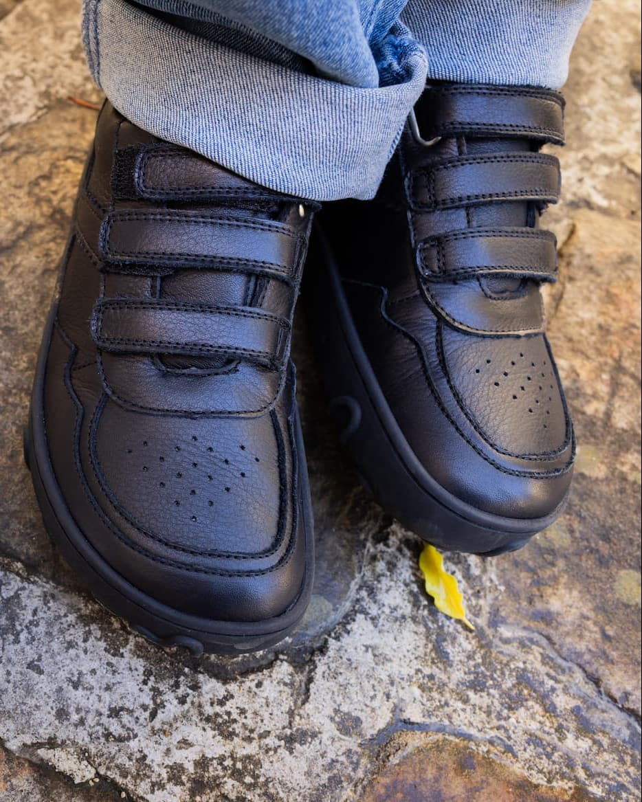 Black leather toddler barefoot sneakers with three hook-and-loop straps, shown on a child's feet with cuffed blue jeans standing on a stone surface.