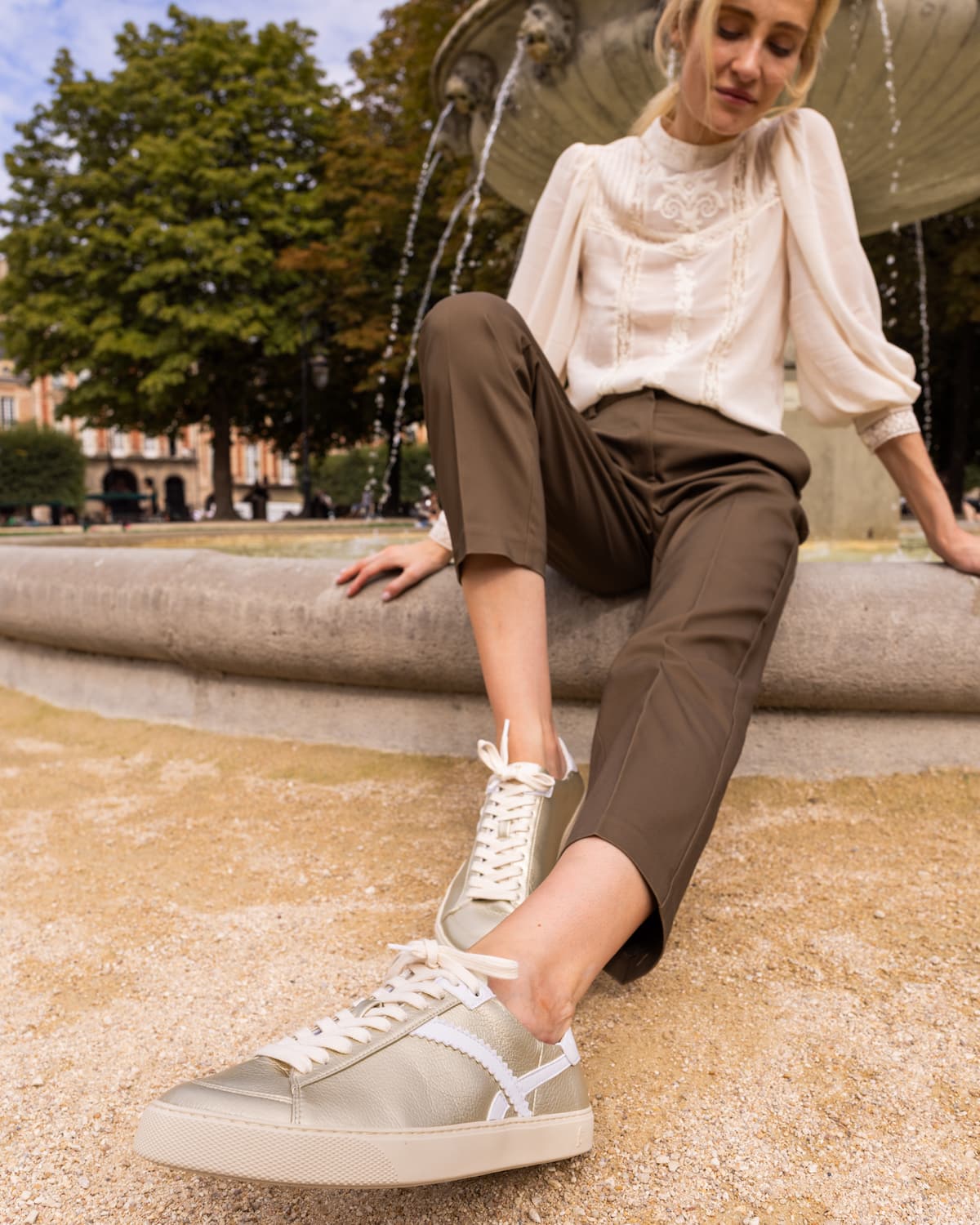 A woman sitting on a stone fountain in an outdoor park setting, wearing a cream blouse, brown cropped trousers, and gold barefoot sneakers with white laces.