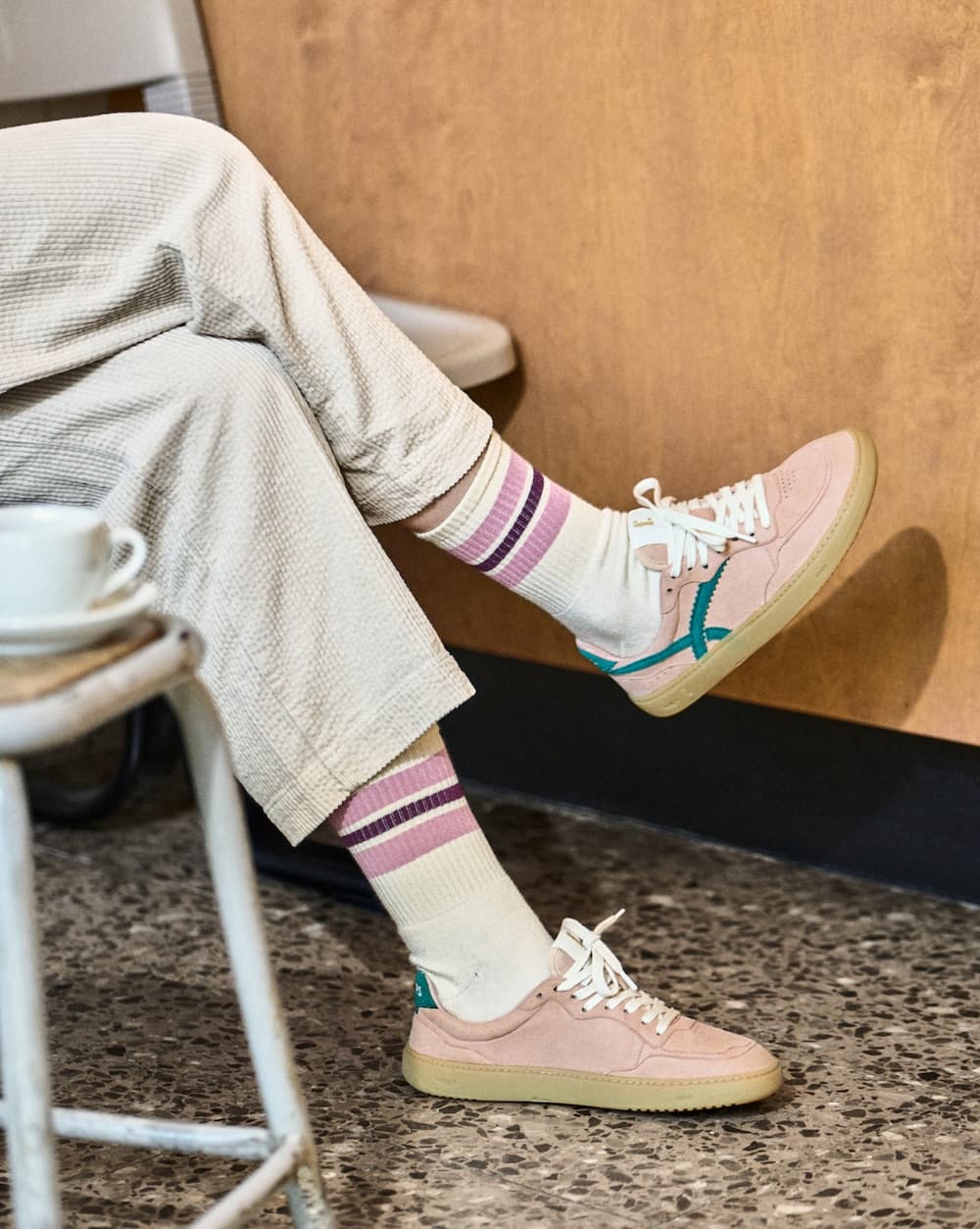 Person wearing light pink barefoot sneakers with green accents, striped socks, and beige pants sitting on a chair.
