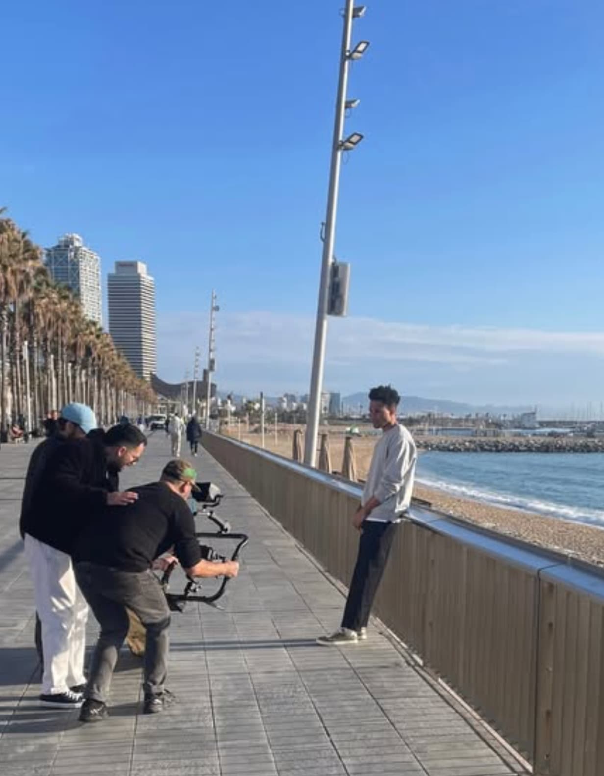 People on a promenade by the sea with buildings and palm trees in the background.