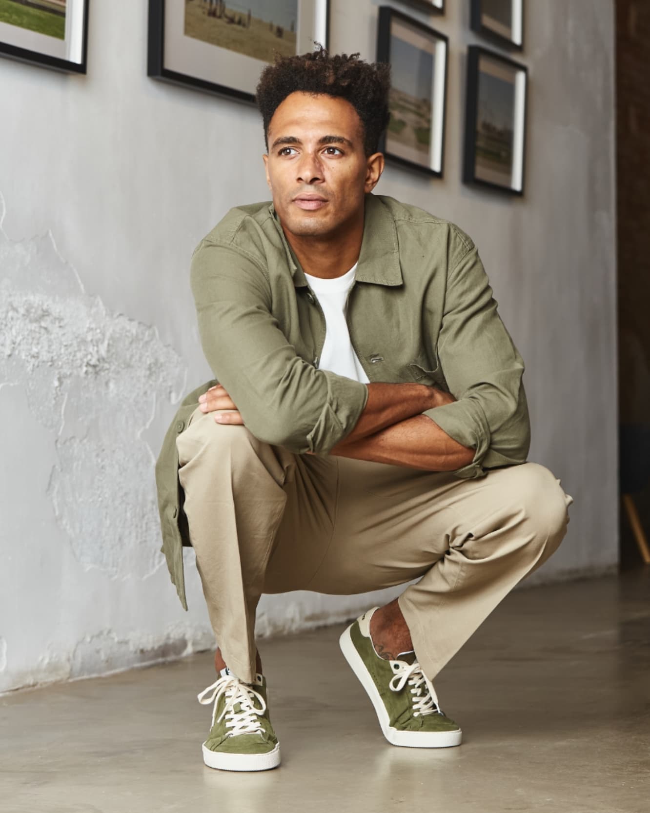 Man in green jacket, khaki pants, green barefoot sneakers sitting on the floor with framed pictures on the wall.