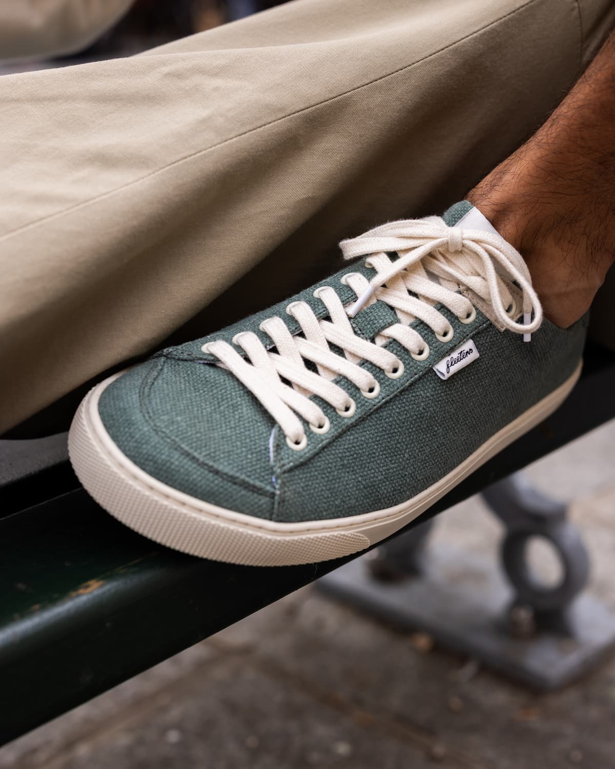 A close-up of a person's foot wearing a green wool barefoot sneaker with white laces and a cream sole, sitting on a dark green bench.